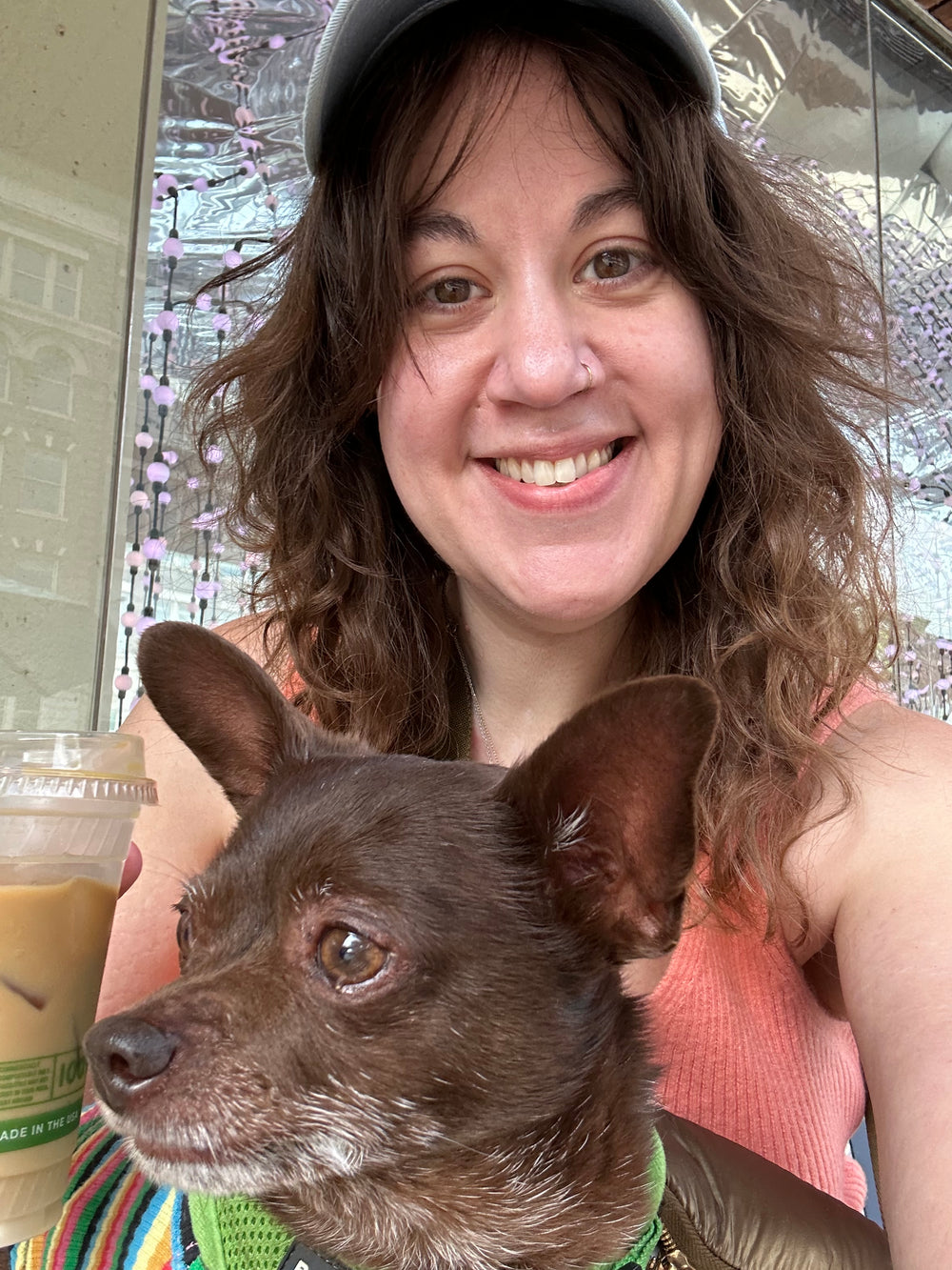 A smiling woman with long wavy brown hair and a cap holds a small dark brown dog in a selfie-style photo. She is wearing a peach-pink ribbed tank top and a colorful striped collar or harness, with an iced coffee in one hand.