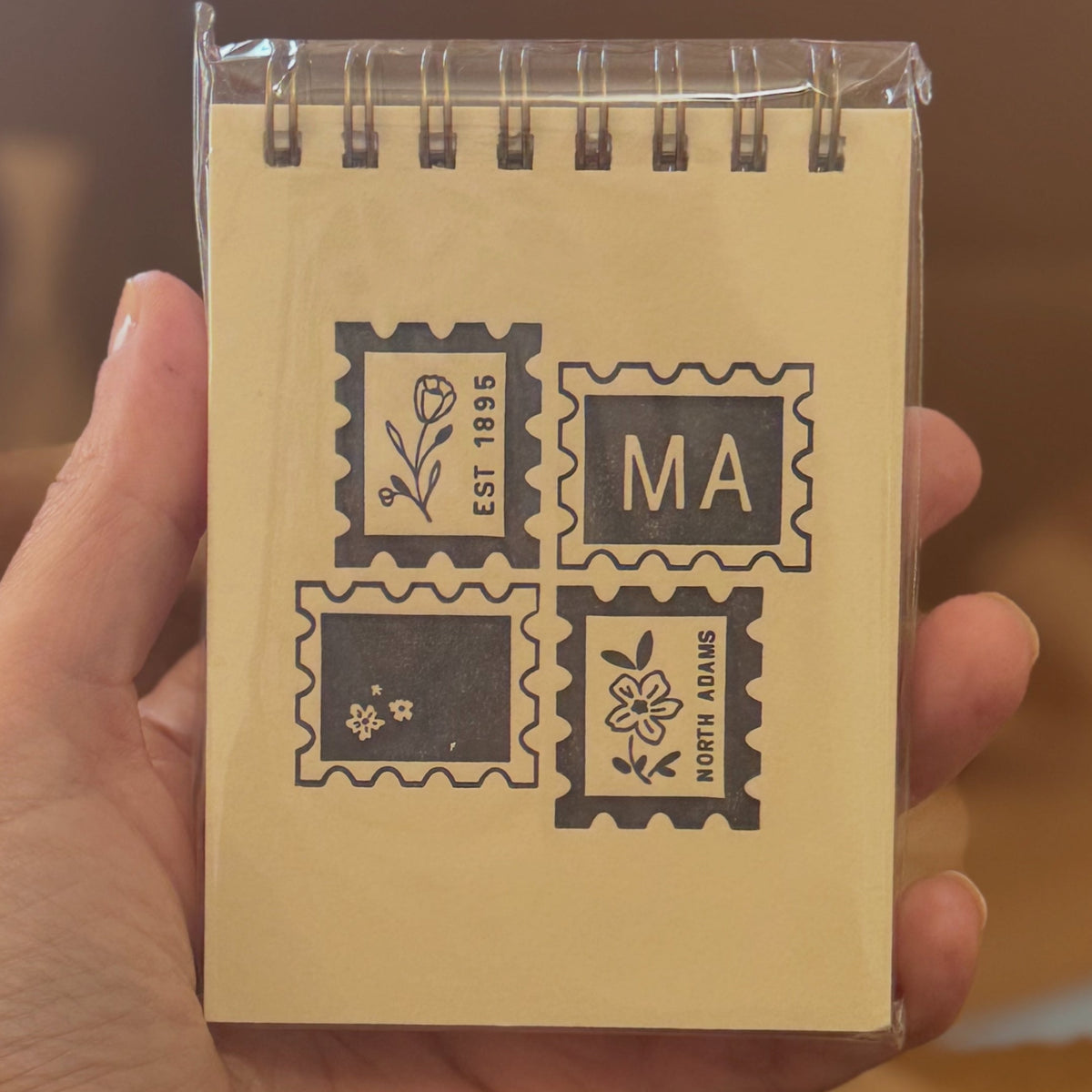 A hand holding a small spiral-bound notebook with a beige cover decorated with four black postage stamp designs, including one that says 'MA' and another with a flower.